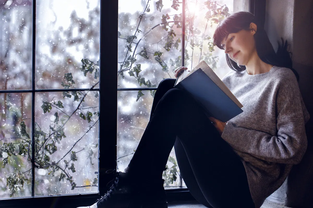 what-to-look-for-in-a-new-window Woman reading by a large insulated window on a snowy day, illustrating what to look for in a new window.